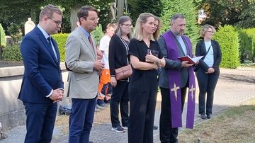 Auf dem Bild sieht man vier Personen in einer Reihe stehen. Man kann erahnen, dass sie vor den Särgen stehen, die man aber nicht sieht. In der Mitte steht Pfarrer Lewinksi, erkennbar an einem Ornat in weiß und violett. Links neben ihm steht, ganz in schwarz gekleidet, Pastoralreferentin Liffers. 