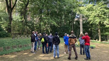 Auf einer Waldlichtung, vor einem hohen Kreuz, umgeben von Grabsteinen  stehen etwa 15 Personen und hören einem Mann zu, der etwas erzählt.