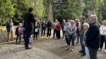 Eine größere Gruppen steht vor einer Steinterrasse und hört einem Mann in schwarzem Anzug zu.