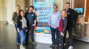 Auf dem Bild sieht man sechs Personen - drei Frauen, drei Männer - rechts und links neben einem Roll-up stehen und in die Kamera lächeln. Auf dem Roll-up steht: "Wenn Lehrkräfte über sich hinaus wachsen".