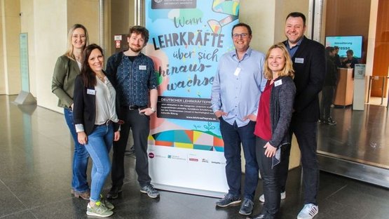 Auf dem Bild sieht man sechs Personen - drei Frauen, drei Männer - rechts und links neben einem Roll-up stehen und in die Kamera lächeln. Auf dem Roll-up steht: "Wenn Lehrkräfte über sich hinaus wachsen".