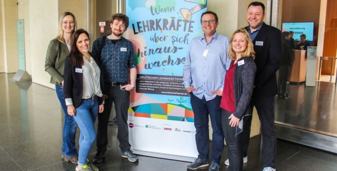 Auf dem Bild sieht man sechs Personen - drei Frauen, drei Männer - rechts und links neben einem Roll-up stehen und in die Kamera lächeln. Auf dem Roll-up steht: "Wenn Lehrkräfte über sich hinaus wachsen".