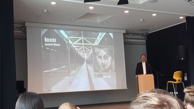 Auf dem Bild sieht man rechts Alwin Meyer hinter einem Redepult und links hinter ihm ein großes Bild aus seiner Präsentation.