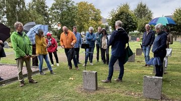 Etwa zwanzig Menschen stehen auf einer Rasenfläche mit eckigen Grabsteinen und hören den Erklärungen eines Mannes zu.