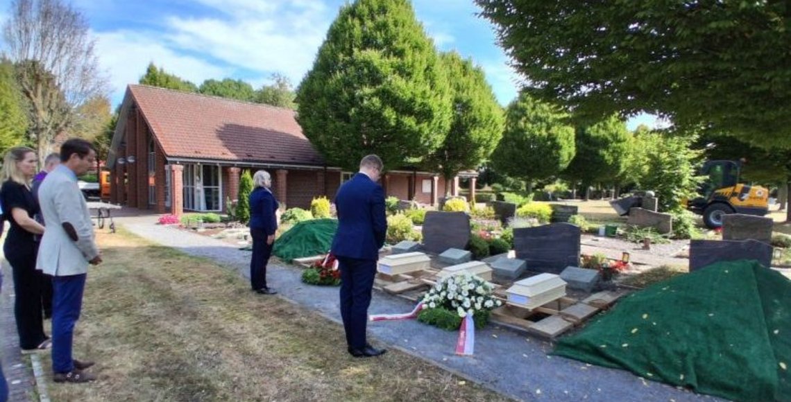 Auf dem Bild sieht man vier Holzsärge vor ausgehobenen Gräbern, dafür stehen die Vertreter des polnischen Generalkonsulats, Frau Wojda und Herr Krzeminski, mit dem gebeugtem Haupt und mit dem Rücken zum Betrachter.