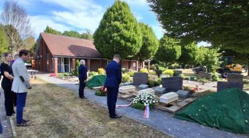 Auf dem Bild sieht man vier Holzsärge vor ausgehobenen Gräbern, dafür stehen die Vertreter des polnischen Generalkonsulats, Frau Wojda und Herr Krzeminski, mit dem gebeugtem Haupt und mit dem Rücken zum Betrachter.