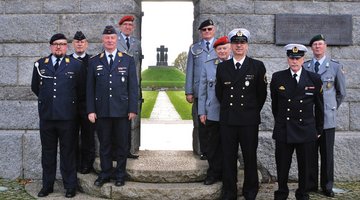 9 Männer in den Uniformen unterschiedlicher Waffengattungen stehen vor einer Mauer, rechts und links eines Tores. Durch das Tor sieht man den Tumulus (Hügel mit Denkmal) der Kriegsgräberstätte La Cambe.