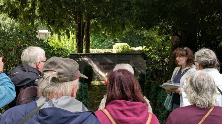 Eine Frau steht vor einem Gedenkstein und erhählt etwas; eine Gruppe von Menschen hört ihr zu.