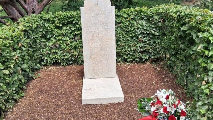 Auf dem Bild sieht man eine Stele, auf der Namen eingraviert sind, umgeben von einer Hecke.