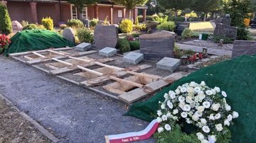 Auf dem Bild sieht man vier ausgehobene Gräber; darauf stehen auf Holzbrettern vier Särge aus hellem Holz. Links und recht liegen jeweils ein Kranz.