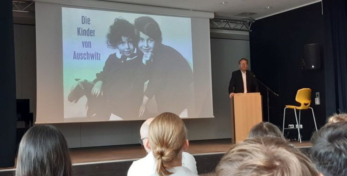 Auif dem Bild sieht man rechts im Bild Alwin Meyer an einem Redepult, hinter him ein großes Bild mit zwei Kinderportraits und der Aufschrift "Die Kinder von Auschwitz", im Vordergrund die Hinterköpfe einiger Schülerinnen und Schüler.