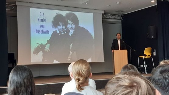 Auif dem Bild sieht man rechts im Bild Alwin Meyer an einem Redepult, hinter him ein großes Bild mit zwei Kinderportraits und der Aufschrift "Die Kinder von Auschwitz", im Vordergrund die Hinterköpfe einiger Schülerinnen und Schüler.