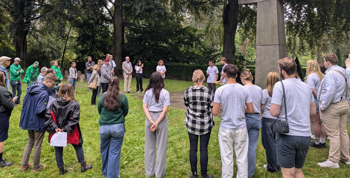 Auf dem Bild sieht man ungefähr 30 vor allem junge Menschen im Halbkreis vor einem überlebensgroßem Steinkreuz stehen.