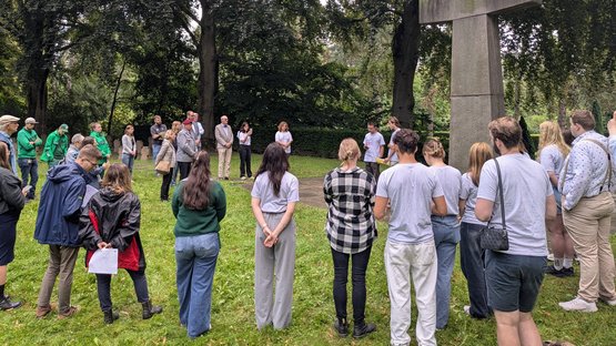 Auf dem Bild sieht man ungefähr 30 vor allem junge Menschen im Halbkreis vor einem überlebensgroßem Steinkreuz stehen.