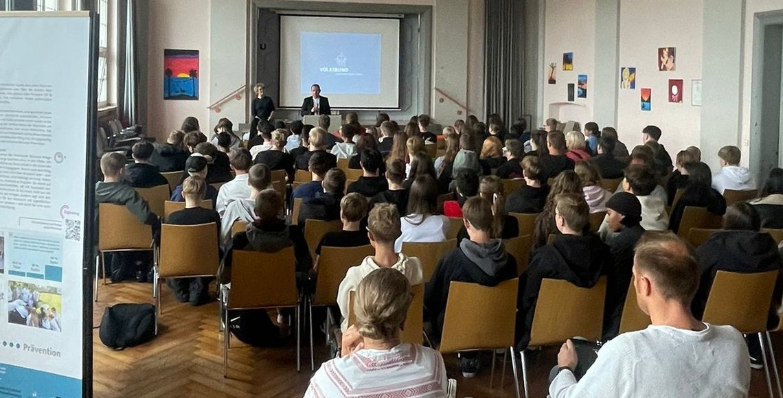 Auf dem Bild blickt man von hinten durch einen Saal über die Köpfe vieler Schülerinnen und Schüler hinweg auf eine Leinwand, vor der ein Mann einen Vortrag hält.